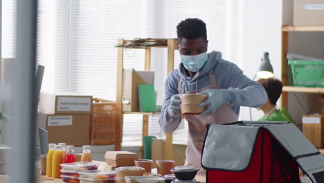Black-Man-in-Mask-Packing-Food-Boxes-and-Drinks-in-Delivery-Bag